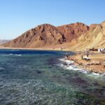 La plage du Blue Hole : les cafés bédouins au pied des montagnes ocre, quelques voitures, la mer agitée au premier plan