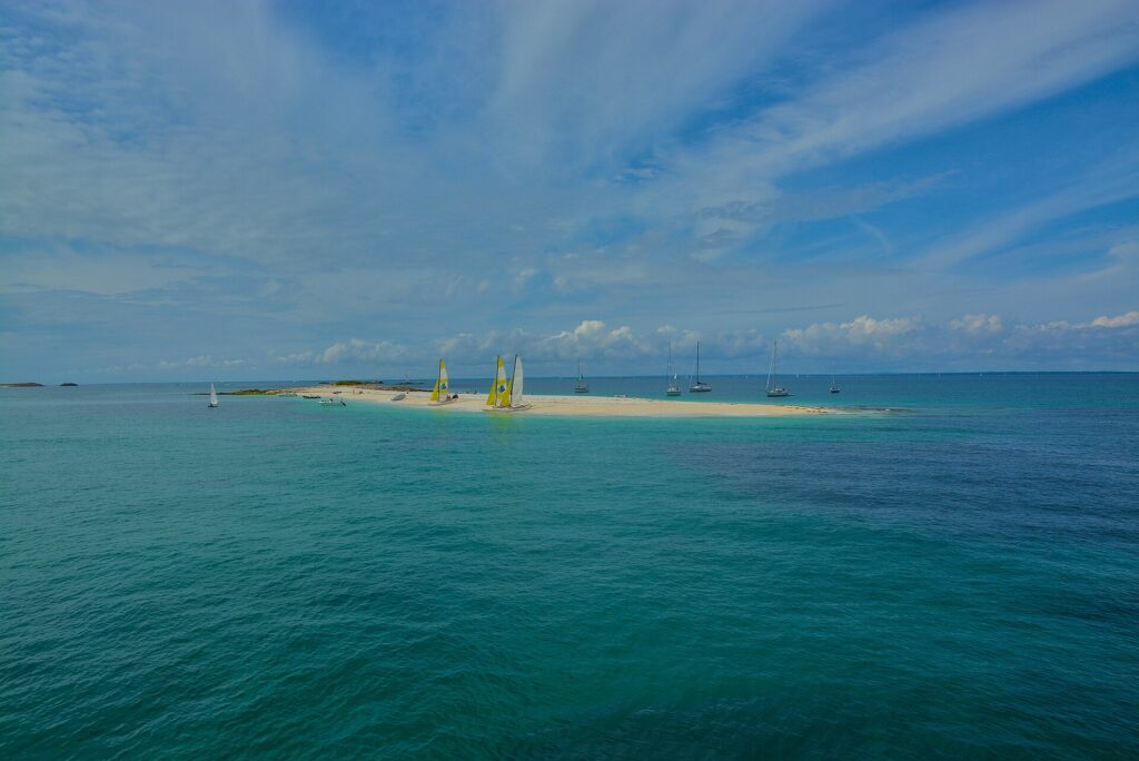 Une longue langue de sable blanc où sont échoués des catamarans aux voiles jaunes et blanches, entourée de voiliers au mouillage sur une eau turquoise, sous un ciel bleu strié de nuages.