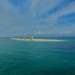 Une longue langue de sable blanc où sont échoués des catamarans aux voiles jaunes et blanches, entourée de voiliers au mouillage sur une eau turquoise, sous un ciel bleu strié de nuages.