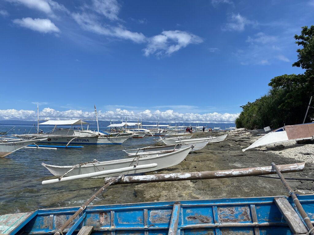 Une file de bangkas blanches amarrées le long d'un rivage rocheux, de petites pirogues à balancier tirées sur la roche et une barque bleue écaillée au premier plan, sous un ciel bleu à nuages blancs.