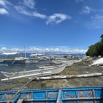 Une file de bangkas blanches amarrées le long d'un rivage rocheux, de petites pirogues à balancier tirées sur la roche et une barque bleue écaillée au premier plan, sous un ciel bleu à nuages blancs.