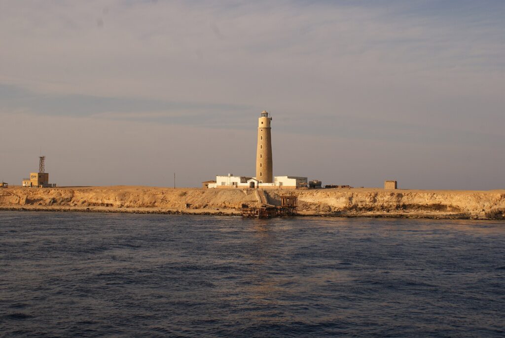 Le phare de Big Brother et ses bâtiments blancs sur l'îlot désertique, une jetée rouillée au premier plan, dans la lumière dorée du soir