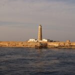Le phare de Big Brother et ses bâtiments blancs sur l'îlot désertique, une jetée rouillée au premier plan, dans la lumière dorée du soir