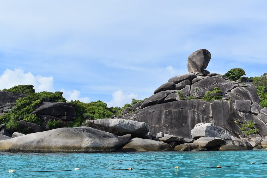 L'énorme bloc de granit en équilibre de l'île 8 coiffe un amas de rochers gris et lisses au bord d'une eau turquoise, sous un ciel bleu voilé, une ligne de bouées au premier plan