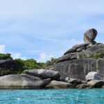 L'énorme bloc de granit en équilibre de l'île 8 coiffe un amas de rochers gris et lisses au bord d'une eau turquoise, sous un ciel bleu voilé, une ligne de bouées au premier plan