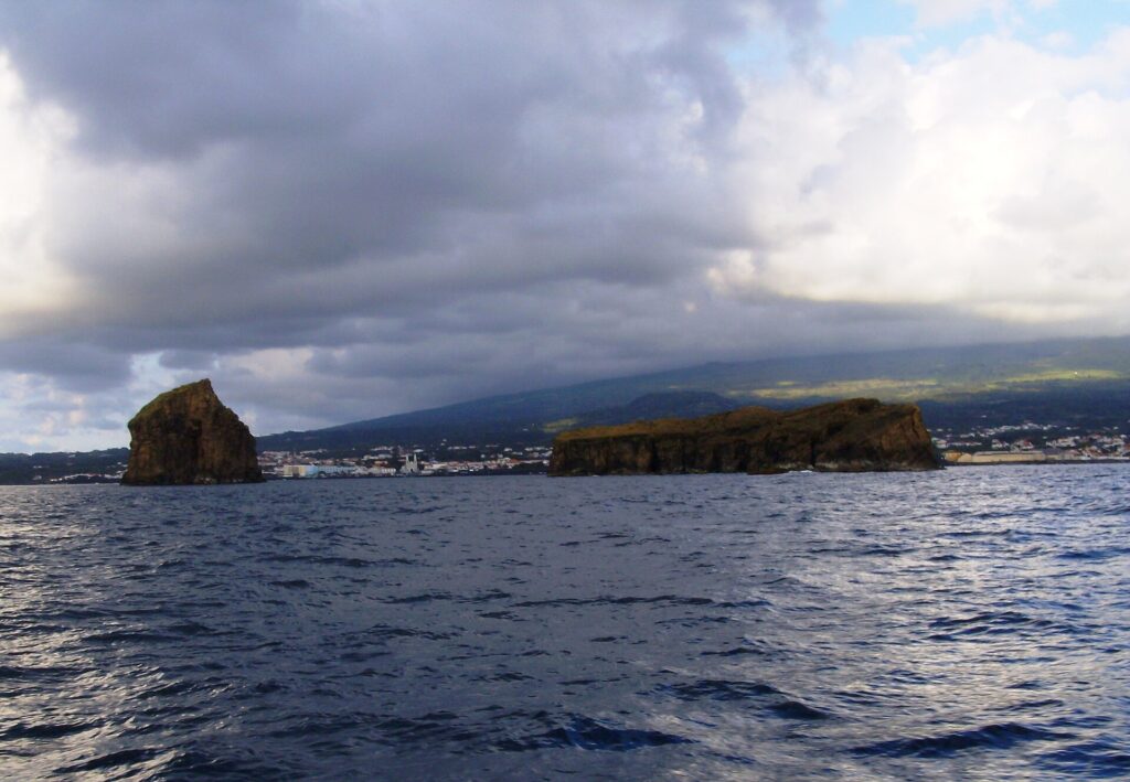 Vus d'un bateau sur une mer sombre et agitée, un îlot rocheux dressé à gauche et un îlot long et plat à droite, devant la ville de Madalena et les pentes de Pico sous un ciel chargé