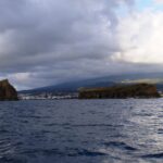 Vus d'un bateau sur une mer sombre et agitée, un îlot rocheux dressé à gauche et un îlot long et plat à droite, devant la ville de Madalena et les pentes de Pico sous un ciel chargé