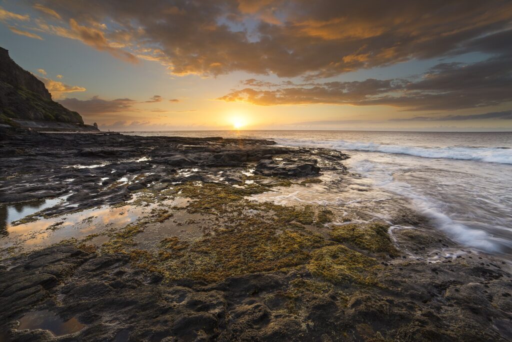 Coucher de soleil sur l'océan vu d'un platier de basalte noir couvert d'algues et de flaques, les vagues qui le lèchent à droite et une falaise à gauche