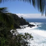Vue plongeante encadrée de palmes : un cap couvert de forêt tropicale plonge dans la mer, les vagues blanchissent sur des rochers noirs et une côte lointaine se dessine à l'horizon sous un ciel bleu.