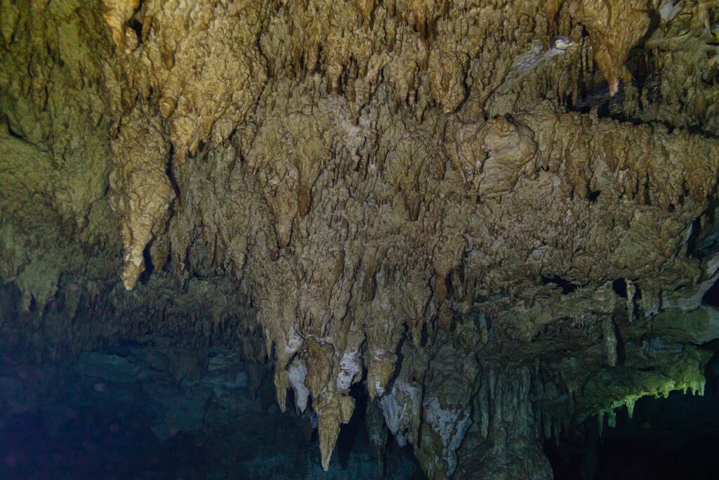 Une forêt de stalactites beiges qui descend vers l'eau sombre, avec une colonne au centre et une lueur verte à droite