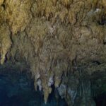 Une forêt de stalactites beiges qui descend vers l'eau sombre, avec une colonne au centre et une lueur verte à droite