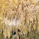 Gros plan sur des concrétions jaune pâle et des stalactites fines, avec le reflet ondulé de l'eau en bas