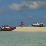 Un banc de sable blanc entouré d'eau claire vert pâle, où un pneumatique noir et orange vient d'accoster, des personnes en gilet de sauvetage sur le sable et la station des gardes sur pilotis à droite.