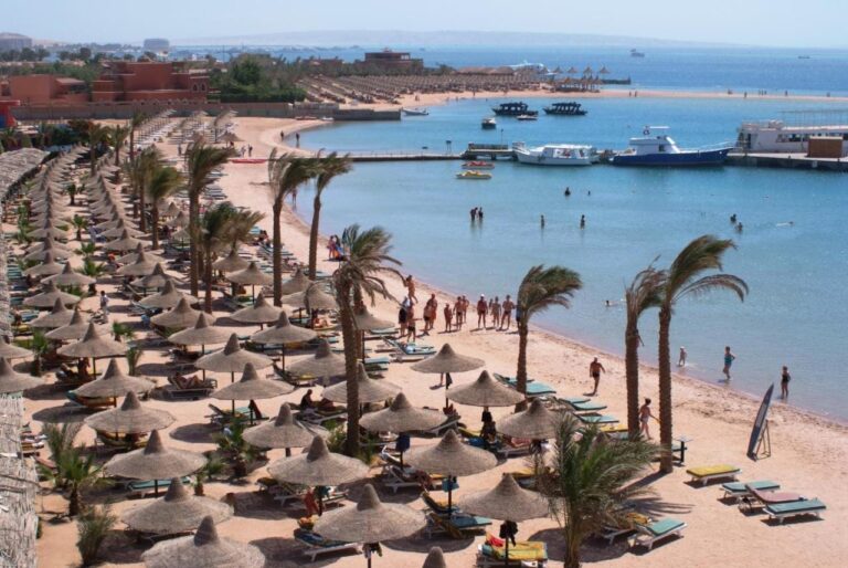 La plage du Giftun Azur Resort, ses parasols de paille et ses palmiers, avec une jetée et des bateaux au fond