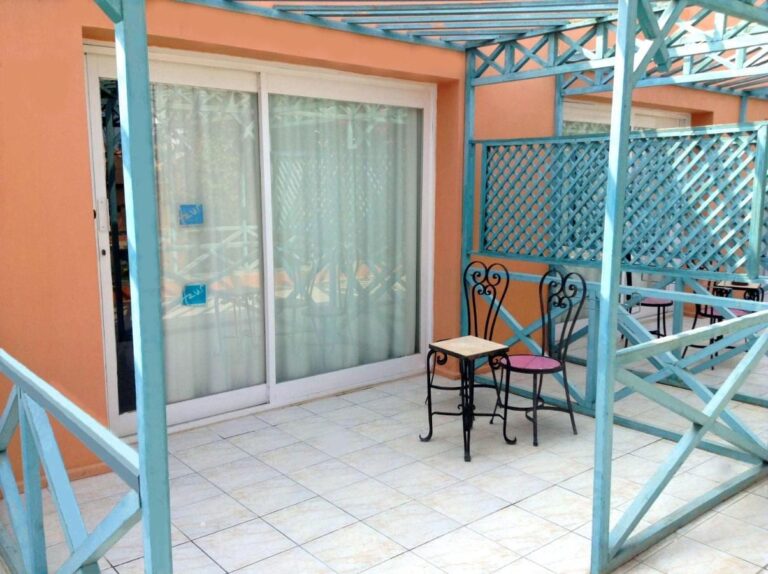 Une terrasse carrelée sous une pergola turquoise, avec une petite table et deux chaises en fer forgé