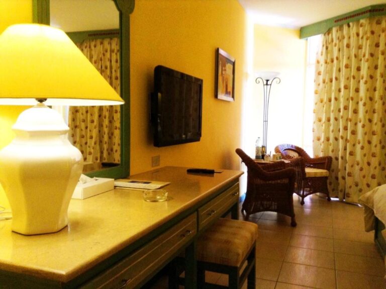 Une chambre aux murs jaunes, avec un bureau, une télévision et deux fauteuils en rotin près de la fenêtre