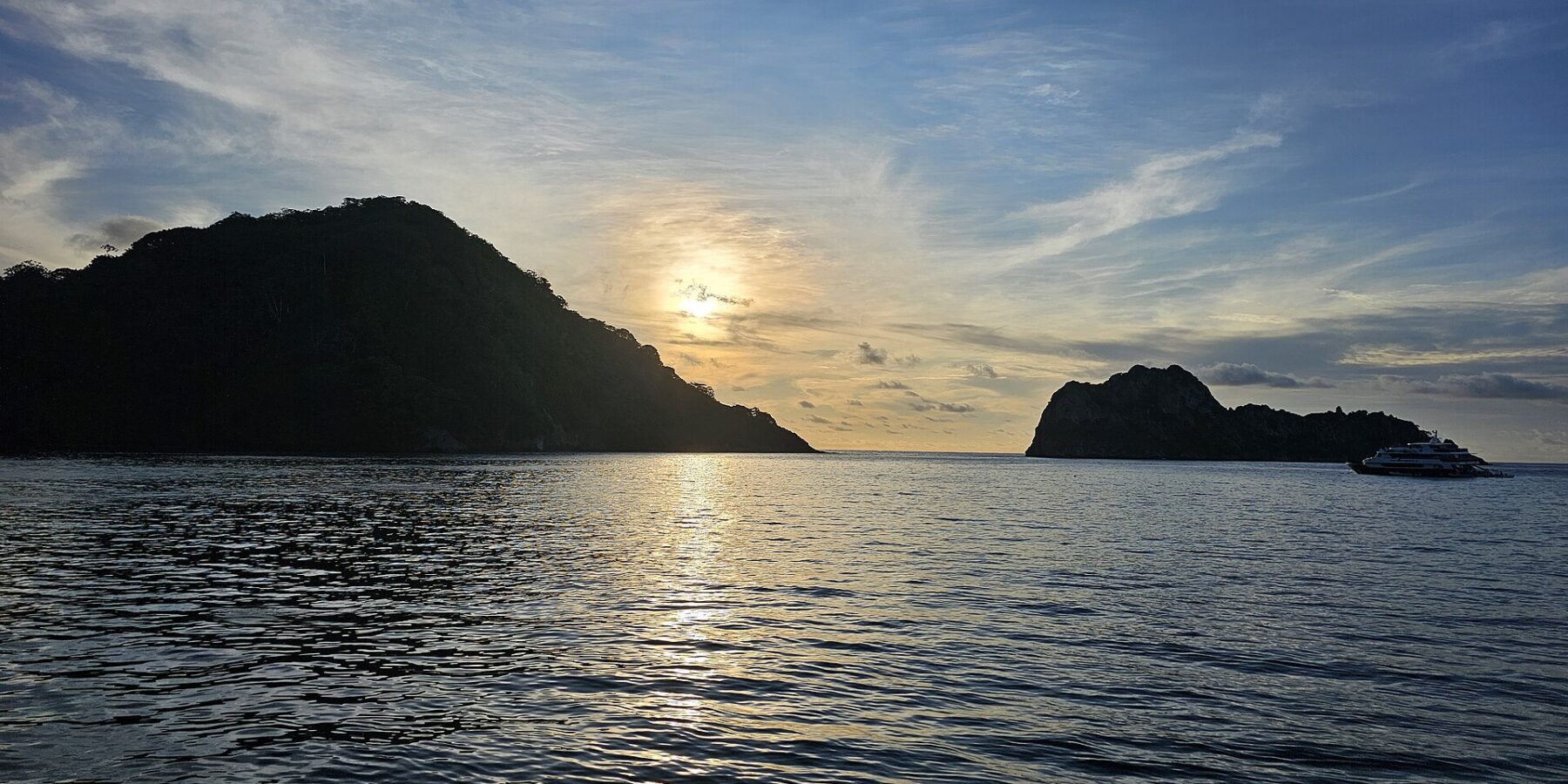 L'île Cocos en contre-jour au coucher du soleil, un bateau de croisière au mouillage à droite, la mer calme