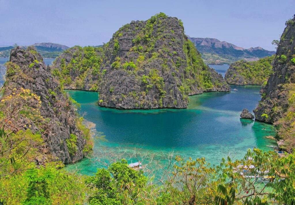 Vue plongeante sur une crique turquoise enserrée de pitons calcaires couverts de végétation, quelques bangkas amarrées en bas et des collines à l'horizon.
