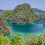 Vue plongeante sur une crique turquoise enserrée de pitons calcaires couverts de végétation, quelques bangkas amarrées en bas et des collines à l'horizon.