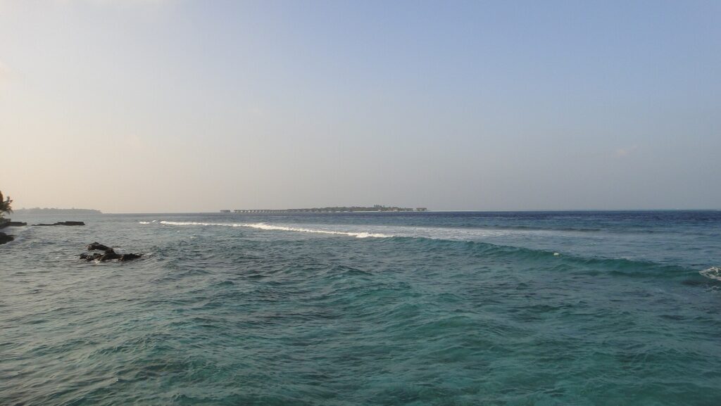 Depuis un rivage rocheux, une passe turquoise agitée où une ligne de vagues déferle sur le récif, et à l'horizon une île basse bordée de bungalows sur pilotis, sous un ciel pâle.