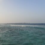 Depuis un rivage rocheux, une passe turquoise agitée où une ligne de vagues déferle sur le récif, et à l'horizon une île basse bordée de bungalows sur pilotis, sous un ciel pâle.