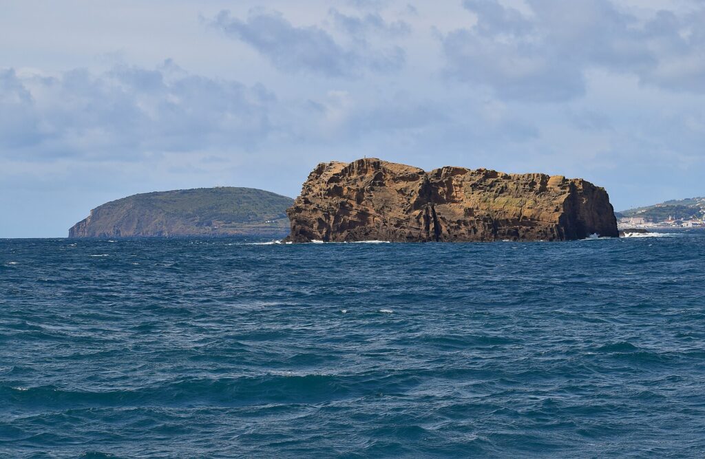 L'Ilhéu Deitado, long bloc de tuf ocre aux falaises creusées d'alvéoles, sur une mer bleu sombre agitée, avec derrière lui la colline verte du Monte da Guia et des maisons de Faial au loin à droite