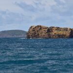 L'Ilhéu Deitado, long bloc de tuf ocre aux falaises creusées d'alvéoles, sur une mer bleu sombre agitée, avec derrière lui la colline verte du Monte da Guia et des maisons de Faial au loin à droite