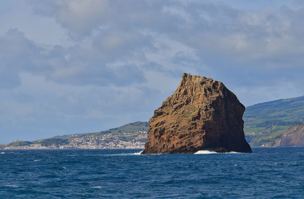 L'Ilhéu em Pé, haut rocher de tuf ocre aux strates creusées, se dresse dans une mer bleu sombre où brisent quelques vagues, devant la ville blanche de Horta et les collines vertes de Faial sous un ciel nuageux