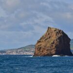 L'Ilhéu em Pé, haut rocher de tuf ocre aux strates creusées, se dresse dans une mer bleu sombre où brisent quelques vagues, devant la ville blanche de Horta et les collines vertes de Faial sous un ciel nuageux