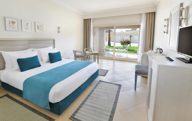 Une chambre au grand lit aux meubles clairs, ouverte sur une terrasse et une pelouse fleurie
