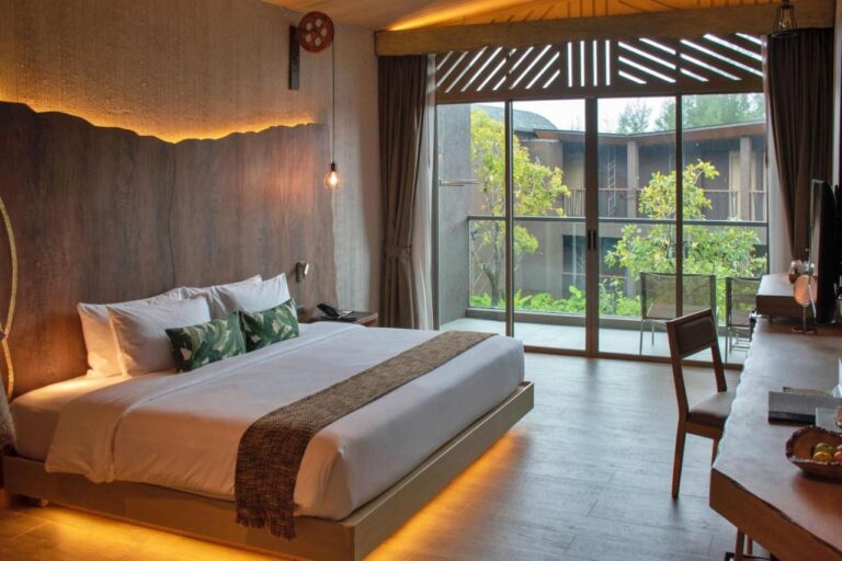 Une chambre au grand lit ouverte sur un balcon, face aux arbres et à un autre bâtiment