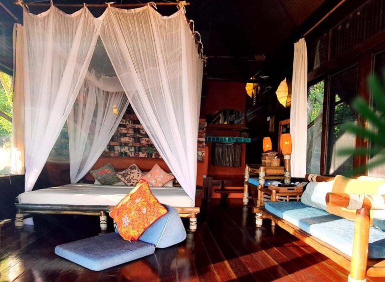 Un cottage avec un lit sous moustiquaire, une banquette en bambou aux coussins bleus et un parquet sombre