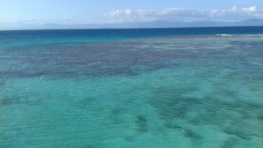 Le lagon vu d'en haut, une eau turquoise tachée de massifs de corail sombres, la barrière où brisent les vagues à droite et les montagnes de la Grande Terre à l'horizon