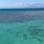 Le lagon vu d'en haut, une eau turquoise tachée de massifs de corail sombres, la barrière où brisent les vagues à droite et les montagnes de la Grande Terre à l'horizon