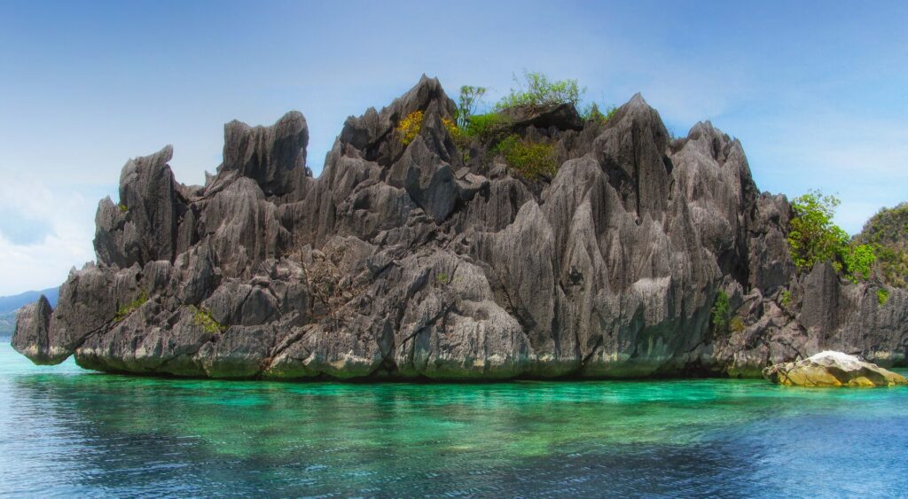 Un îlot de calcaire gris hérissé de pointes, coiffé d'un peu de végétation, se dresse au-dessus d'une eau turquoise transparente où l'on devine le corail, sous un ciel bleu.