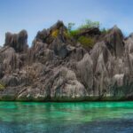 Un îlot de calcaire gris hérissé de pointes, coiffé d'un peu de végétation, se dresse au-dessus d'une eau turquoise transparente où l'on devine le corail, sous un ciel bleu.