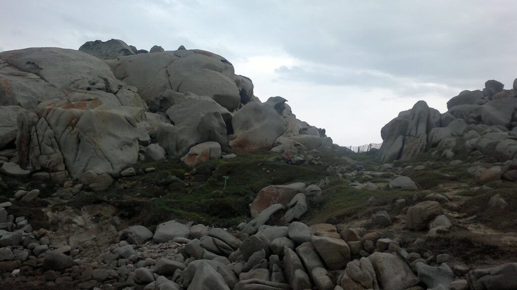 Amas de gros blocs de granite gris arrondis et creusés par l'érosion, tachés de lichens orange, autour d'une pelouse rase où un sentier bordé d'une clôture en bois file vers la mer, sous un ciel couvert.
