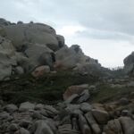 Amas de gros blocs de granite gris arrondis et creusés par l'érosion, tachés de lichens orange, autour d'une pelouse rase où un sentier bordé d'une clôture en bois file vers la mer, sous un ciel couvert.