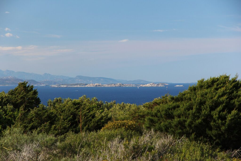 Vue depuis le maquis de la côte sur une longue île basse faite de blocs de granite clair, posée sur une mer bleu sombre, un voilier à ses pieds et des montagnes bleutées à l'horizon.