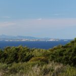 Vue depuis le maquis de la côte sur une longue île basse faite de blocs de granite clair, posée sur une mer bleu sombre, un voilier à ses pieds et des montagnes bleutées à l'horizon.