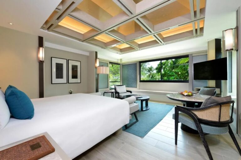 Une chambre au grand lit avec un plafond à caissons, un coin salon et une fenêtre sur les palmiers