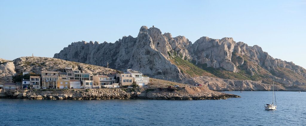 Les maisons du village des Goudes alignées au ras d'une côte de rochers, devant une haute crête de calcaire blanc aux pics déchiquetés, sous un ciel bleu, un voilier au mouillage sur une mer calme.