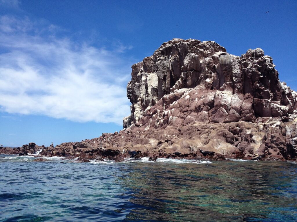 Les rochers roux de Los Islotes sous un ciel bleu, avec des otaries couchées au ras de l'eau et une falaise crevassée au-dessus
