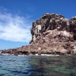 Les rochers roux de Los Islotes sous un ciel bleu, avec des otaries couchées au ras de l'eau et une falaise crevassée au-dessus