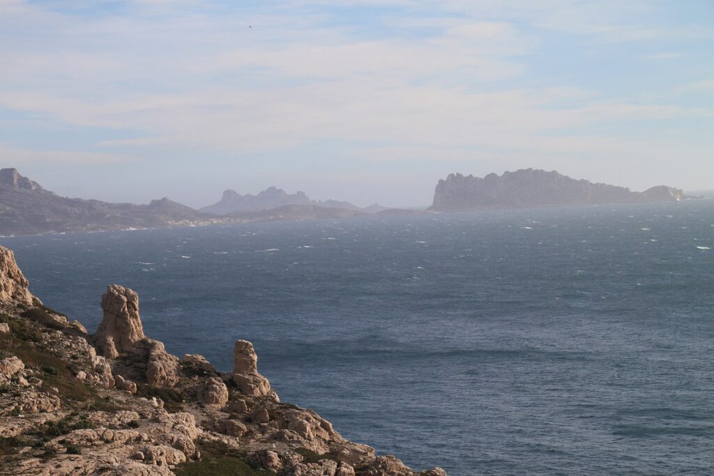Depuis les rochers calcaires du Frioul au premier plan, vue sur une mer agitée et moutonnante jusqu'à l'île Maïre et la côte des Goudes, découpées dans la brume.