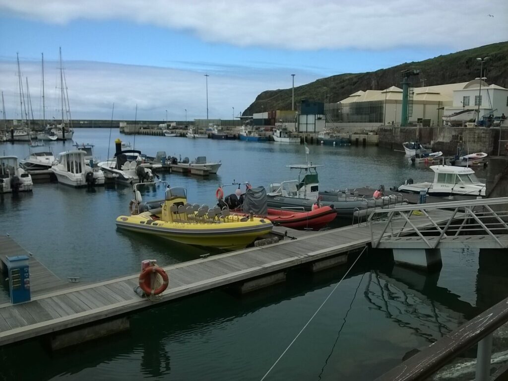 Des semi-rigides, un jaune aux rangées de sièges et un rouge, amarrés au ponton de la marina de Vila do Porto, voiliers et bateaux de pêche au fond sous une falaise verte et un ciel nuageux