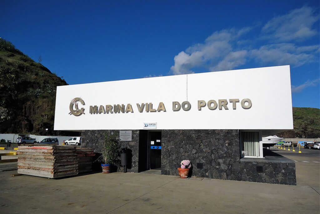 Le bâtiment d'accueil de la marina de Vila do Porto, grande enseigne blanche à lettres métalliques posée sur un mur de basalte noir, sous un ciel bleu, une falaise verte à gauche