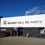 Le bâtiment d'accueil de la marina de Vila do Porto, grande enseigne blanche à lettres métalliques posée sur un mur de basalte noir, sous un ciel bleu, une falaise verte à gauche