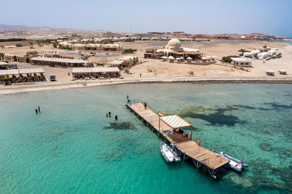 Vue aérienne du Marsa Shagra Village, avec sa jetée, deux zodiacs et le village au bord de l'eau turquoise