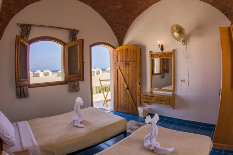 Une chambre à deux lits ouverte sur la plage, avec des tentes blanches et la mer au fond